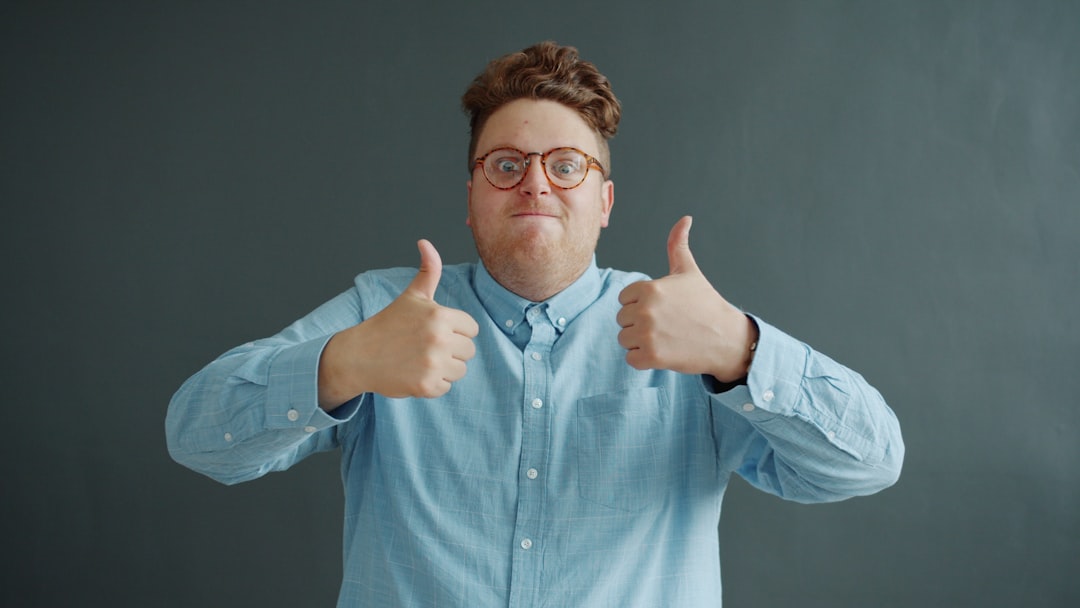 Pleased young man showing thumbs-up hand gesture with both arms on dark gray background standing alone expressing approval. People and lifestyle concept.
